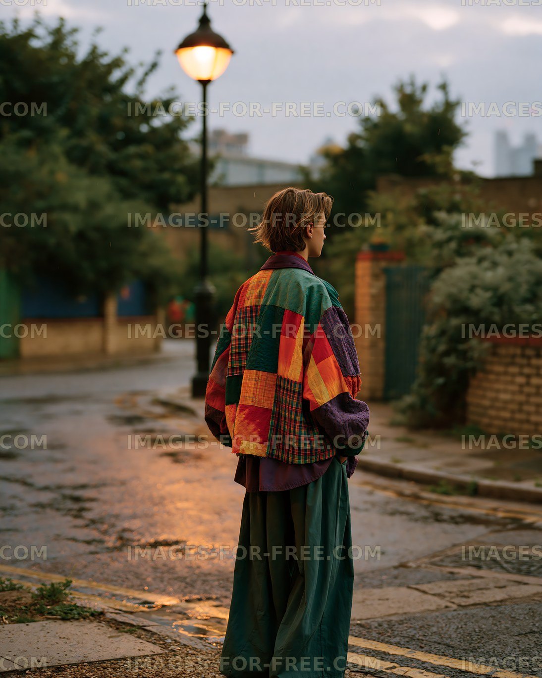 Person wearing colorful clothing stands on a wet street at dusk